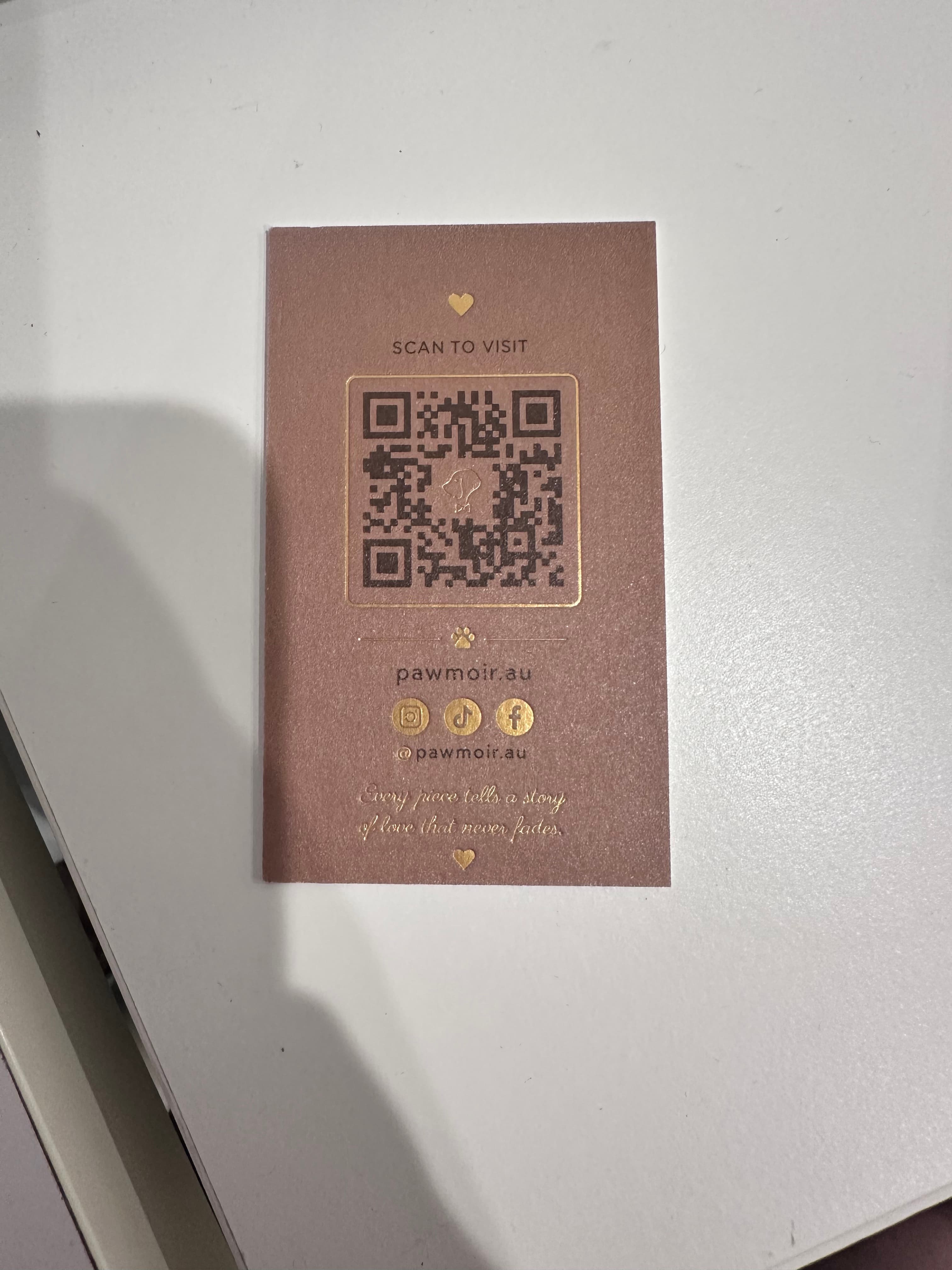 Photograph of the printed Pawmoir Back insert card on a white surface, showing the brown card, gold-toned details, QR graphic, pawmoir.au and social icons.